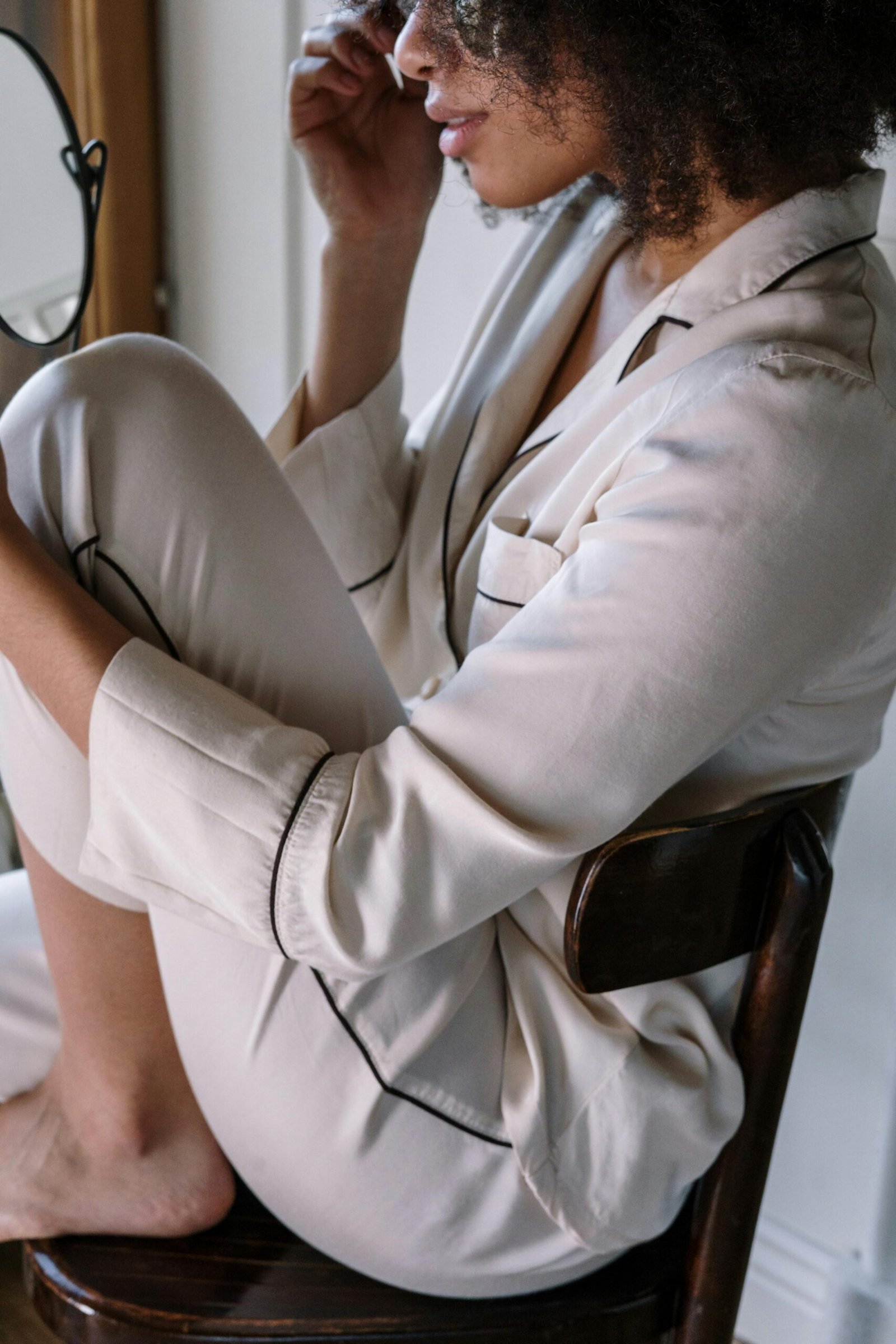 Woman sitting sideways on a wooden chair, wearing cream-colored satin pajamas with black piping, touching her face while looking in a mirror.

