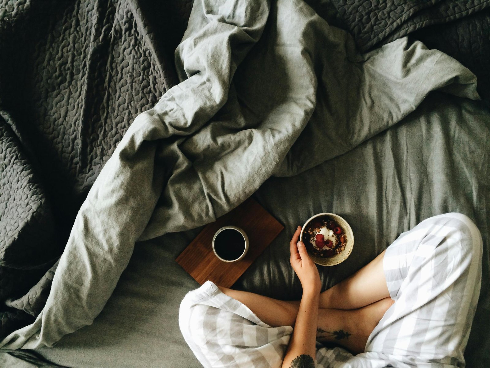 Person sitting cross-legged on a bed with gray bedding, wearing striped pajamas, holding a bowl of food next to a cup of coffee on a wooden tray.

