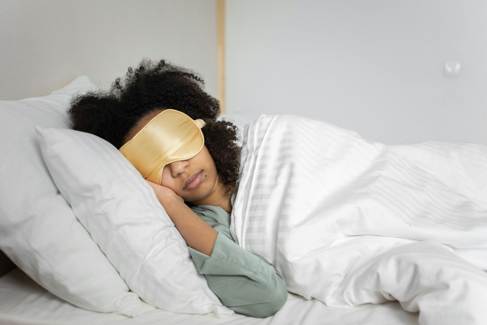 A woman sleeping peacefully in a well-lit room, wearing a gold satin sleep mask and lying under a white blanket on a white bed.

